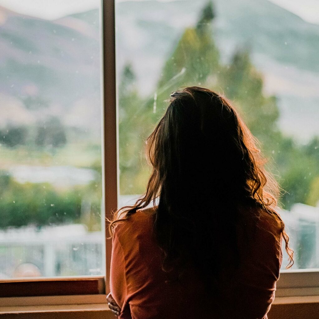 Femme adulte qui regarde le paysage par la fenêtre. Sa silhouette sombre et fermée qui se découpe sur le paysage est emplie de tristesse.